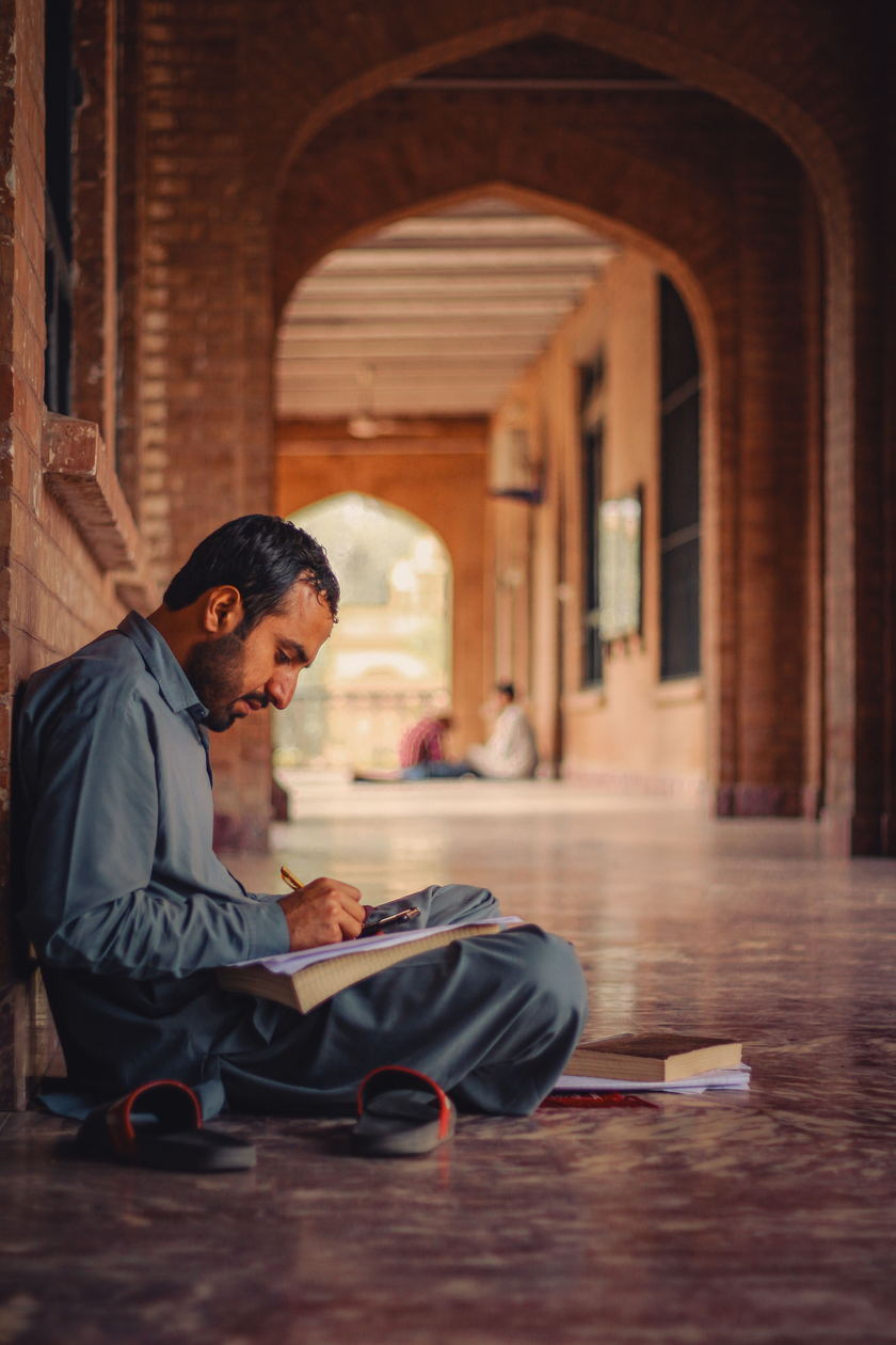 A person writing in a notebook while seated along an arched stone corridor.